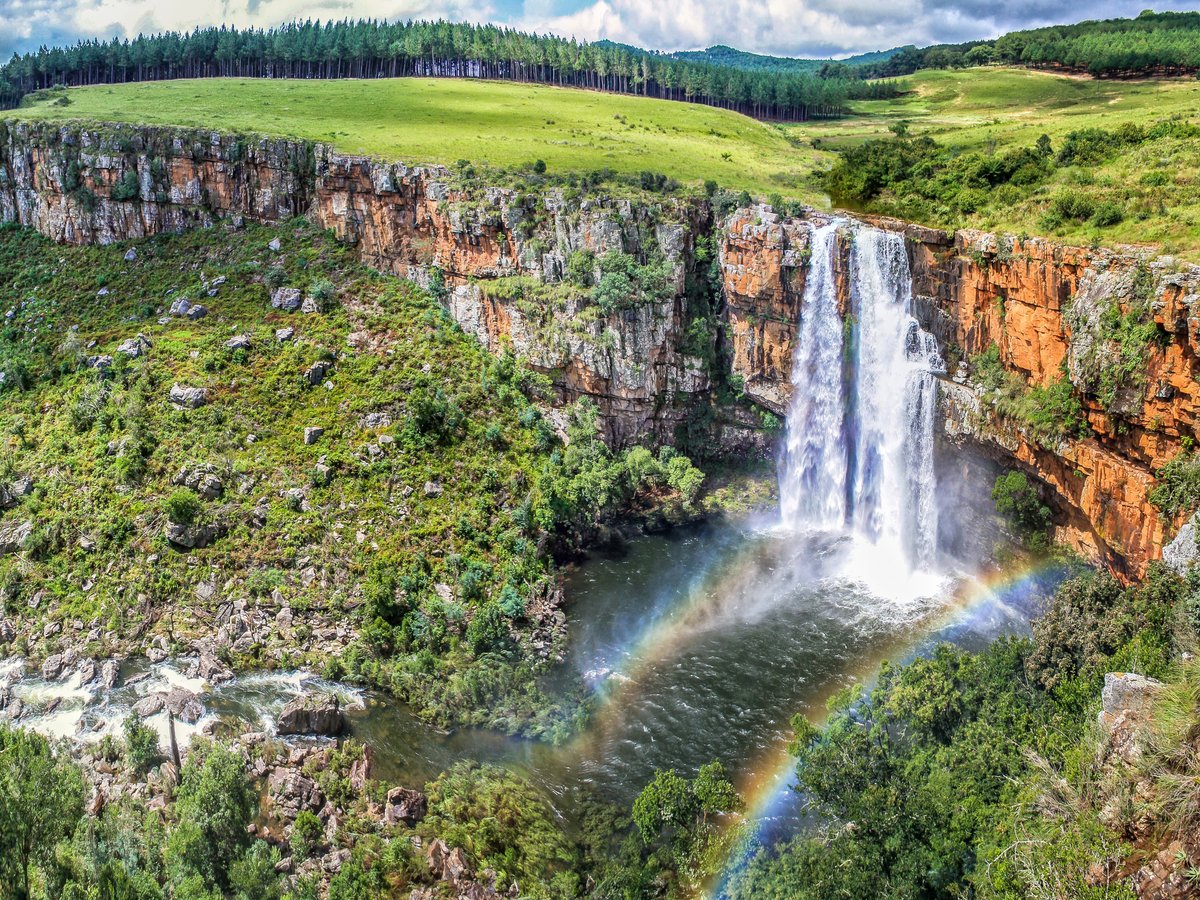 Wasserfall stürzt von einer Felskante in ein bewaldetes Flusstal, über dem Wasser bildet sich ein Regenbogen.