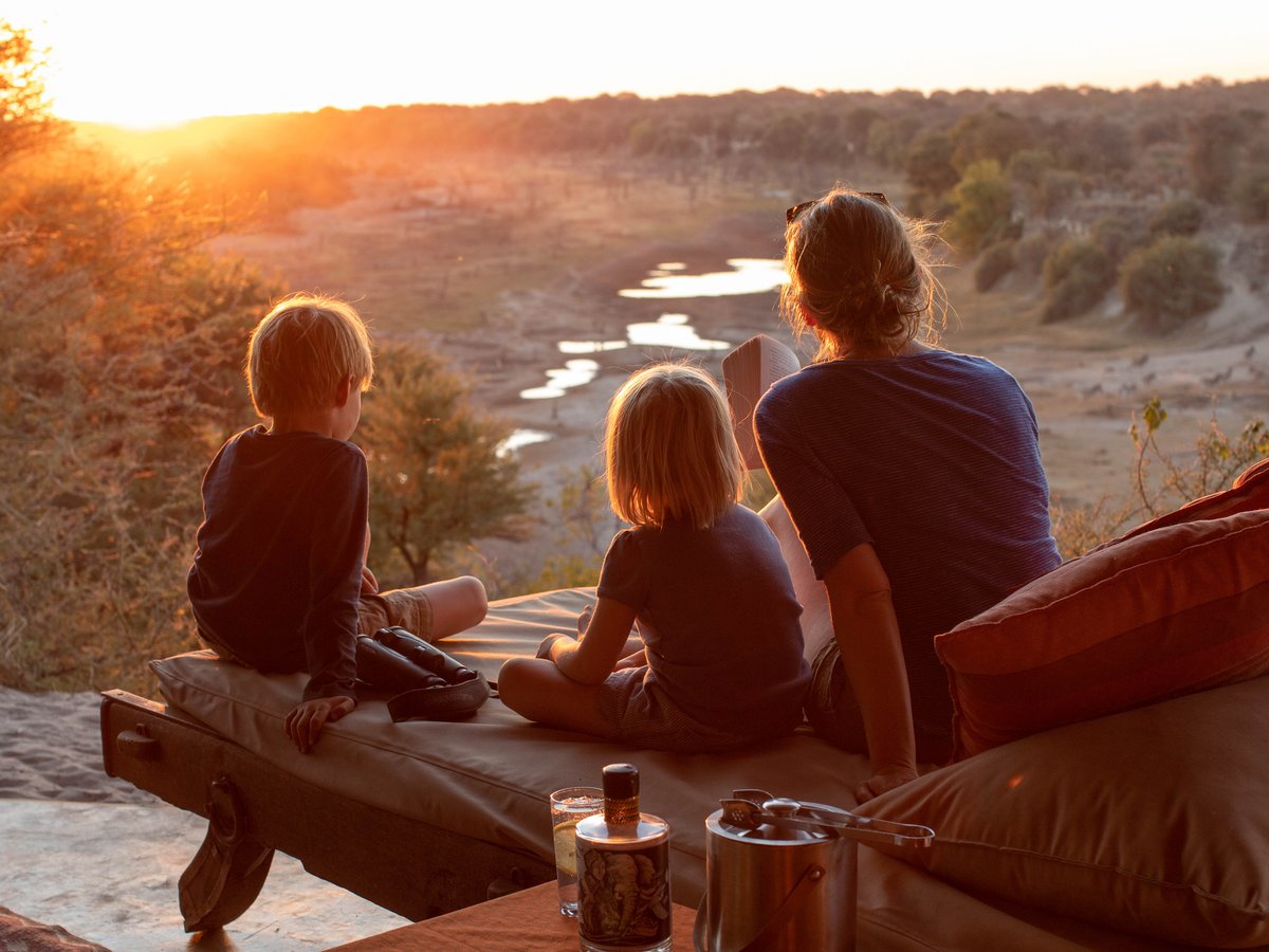 Eine Frau und zwei Kinder sitzen auf einer Liege und blicken auf eine Flusslandschaft bei Sonnenuntergang