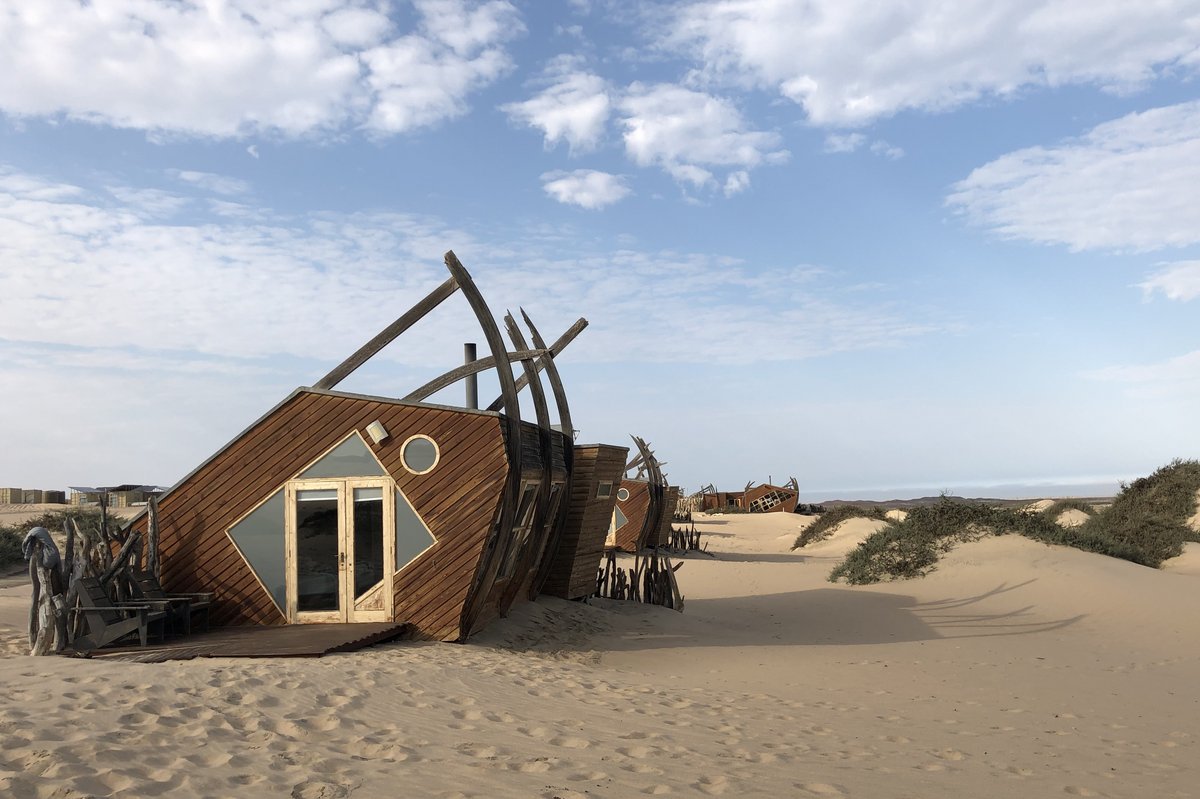 Verwittertes Schiffswrack steht im Sand einer weiten Dünenlandschaft unter Himmel mit weißen Wolken.