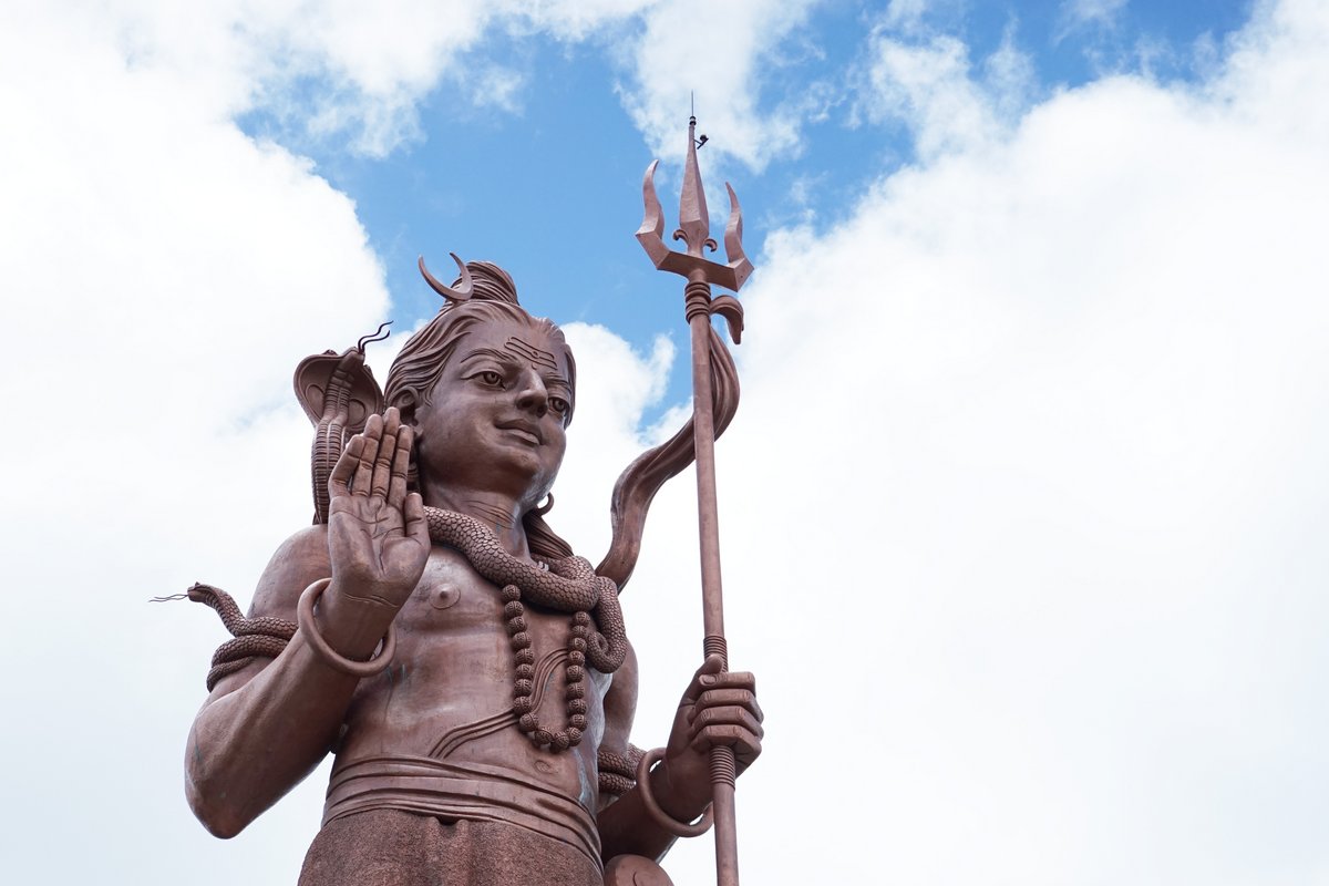 Große Statue einer stehenden Figur mit erhobener Hand, Halskette und Dreizack vor bewölktem Himmel.