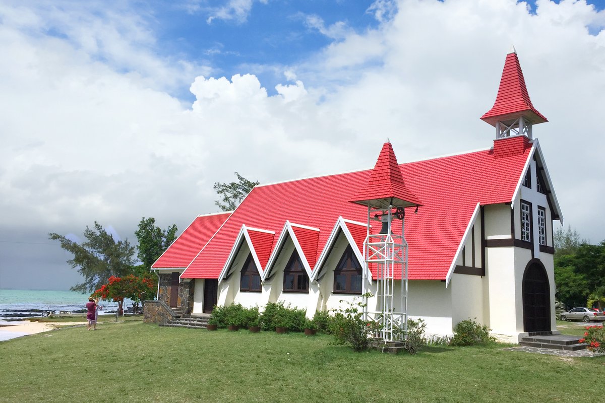 Kirche mit roten Dächern und weißem Fachwerk an einem Strand mit grünem Rasen und bewölktem Himmel.