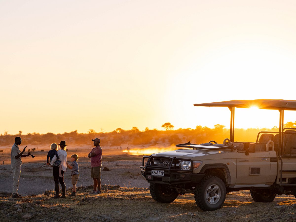 Safari-Geländewagen mit offenem Verdeck steht in der Savanne bei Sonnenuntergang, daneben vier Personen im Gespräch