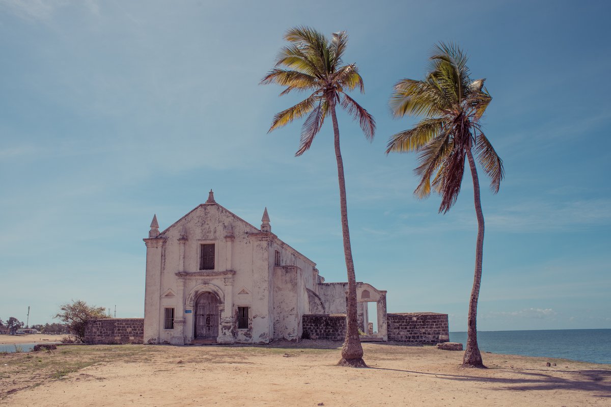 Alte Kirche mit Verzierungen auf einer sandigen Küstenfläche neben drei hohen Palmen und dem Meer im Hintergrund