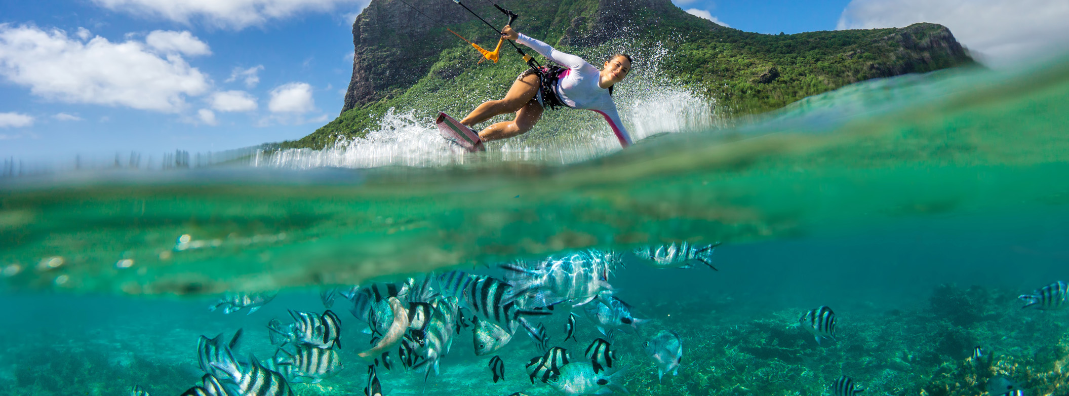 Dynamische Kitsurferin vor prägnantem Berg mit gleichzeitigem Blick auf Fischen unter Wasser. 